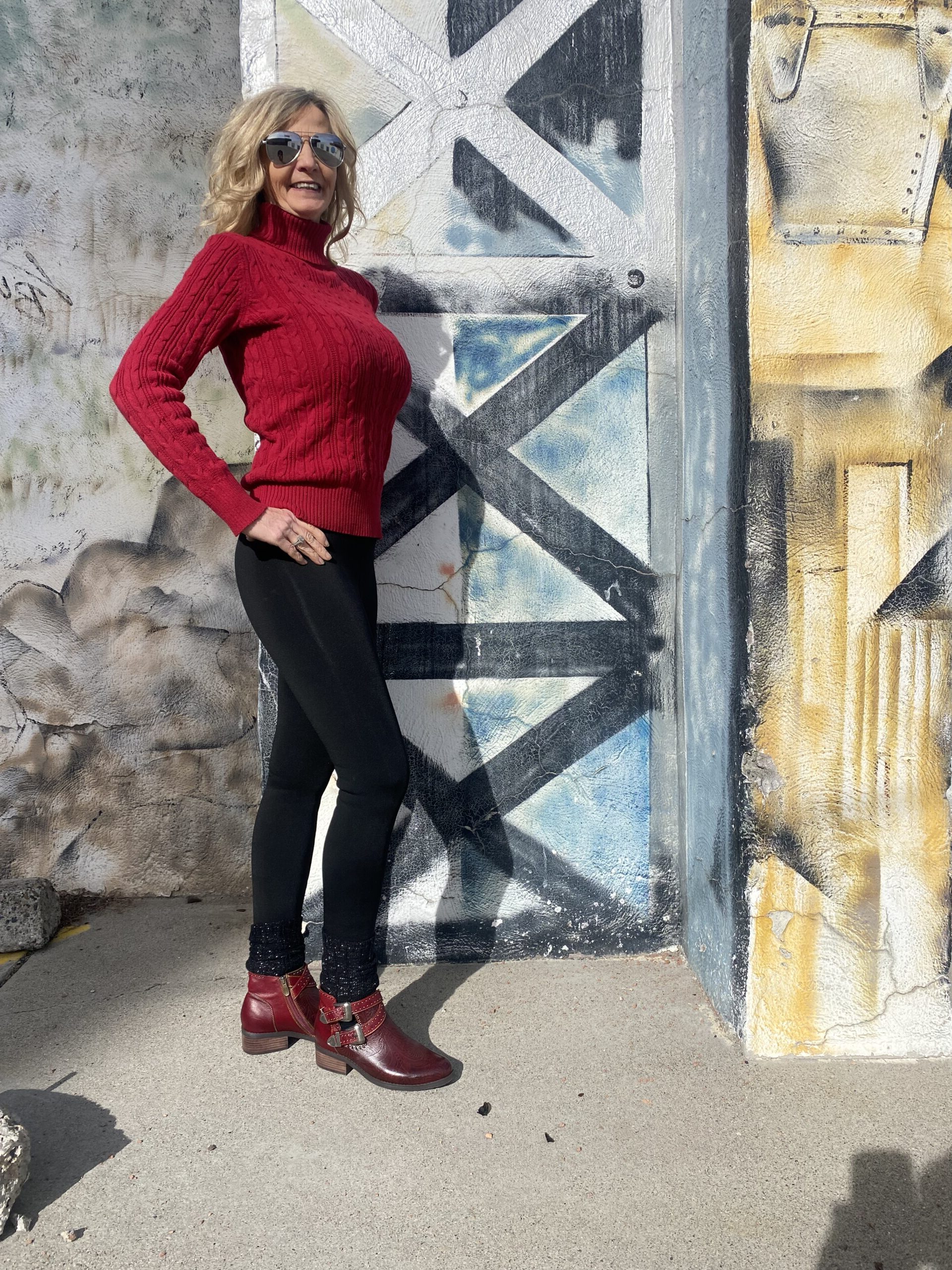 A woman in red sweater and black pants standing next to wall.